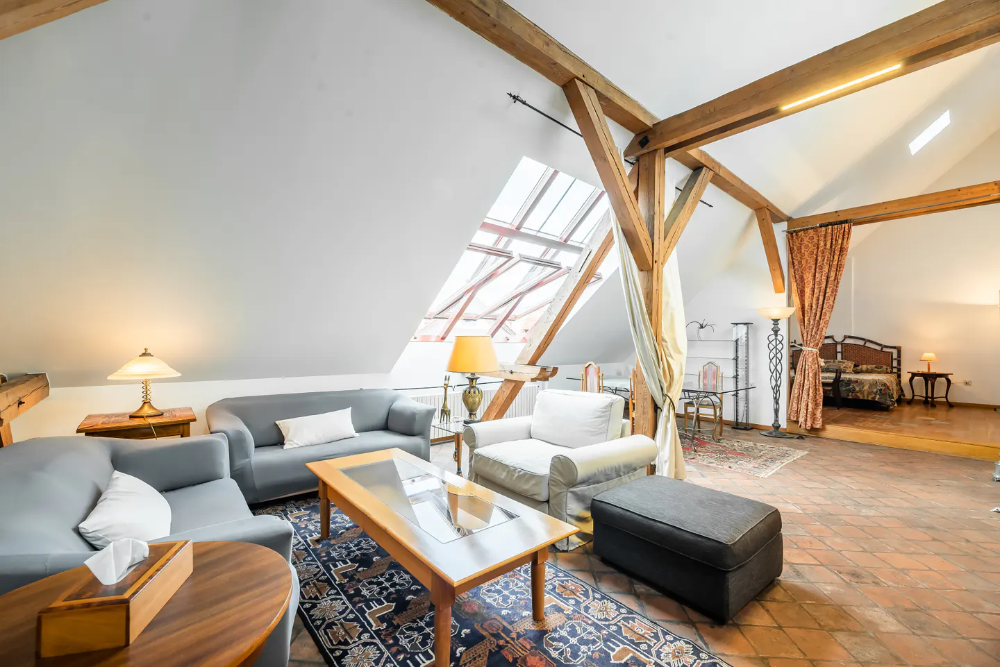 Attic living room with exposed wooden beams, gray sofas, a coffee table, and a raised bedroom area with a patterned curtain.