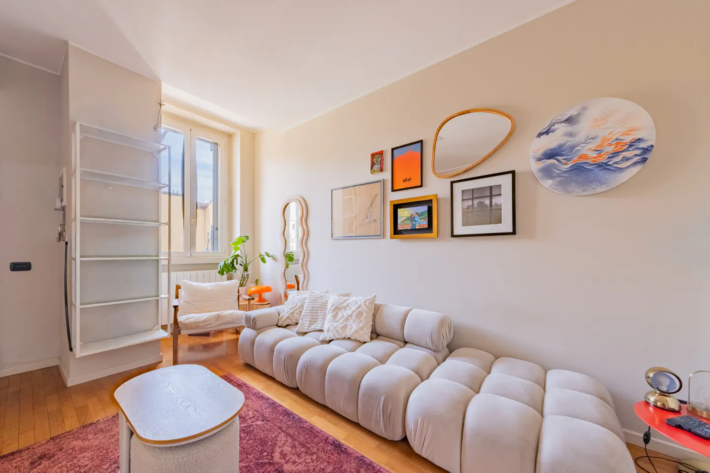 Bright living room with a beige modular sofa, gallery wall, and a purple rug on a wood floor. A white shelving unit is on the left.