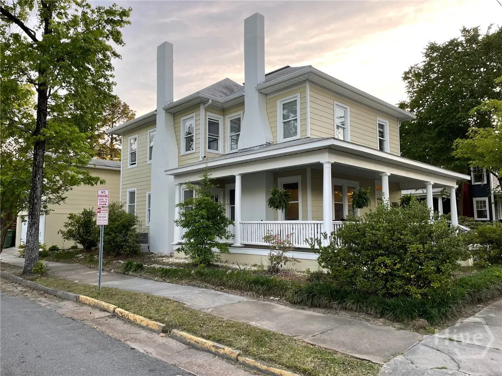 Two-story yellow house with white trim, a porch with columns, and two tall white chimneys. Bushes and trees surround the house.