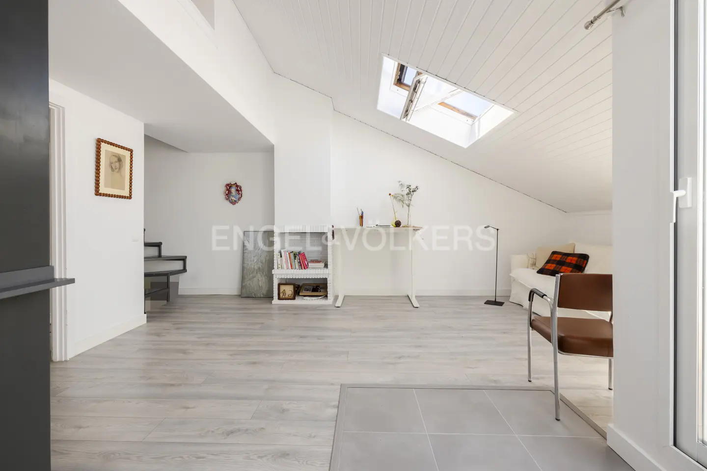 Bright attic room with white walls, slanted ceiling, skylight, light wood floors, a desk, and a sofa with a plaid pillow.