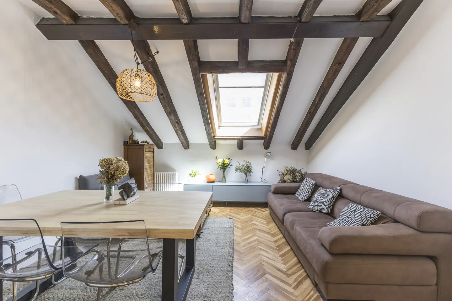 Attic living room with exposed beams, skylight, brown sofa, wood table, and clear chairs on a herringbone wood floor.