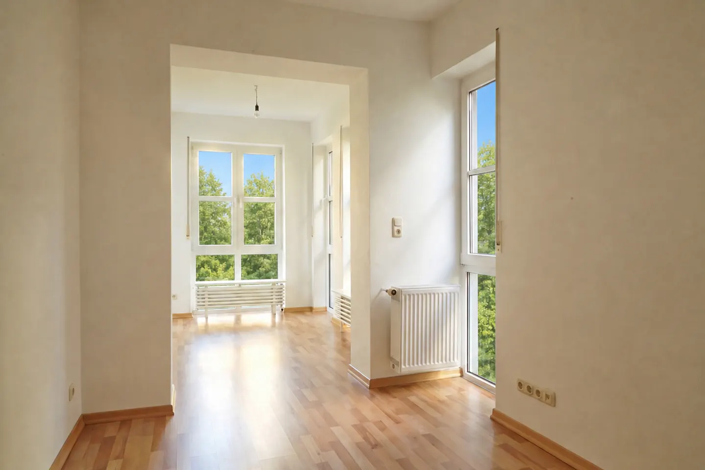 Empty room with light wood floors, white walls, large windows overlooking trees, and white radiators.