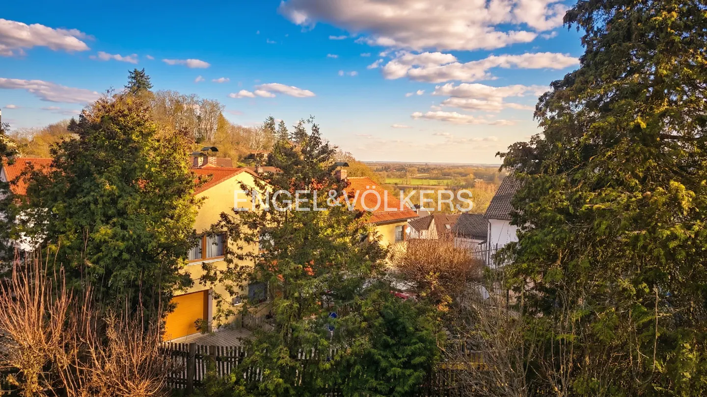 View of yellow houses with red roofs, surrounded by trees, under a blue sky with white clouds. Engel & Völkers logo visible.