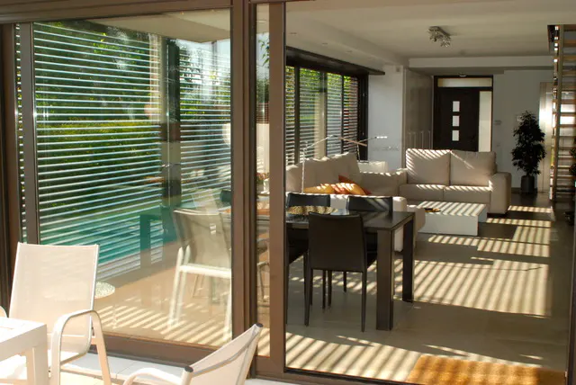 View of a modern living room with a dining table and white sofas, seen through sliding glass doors with blinds.