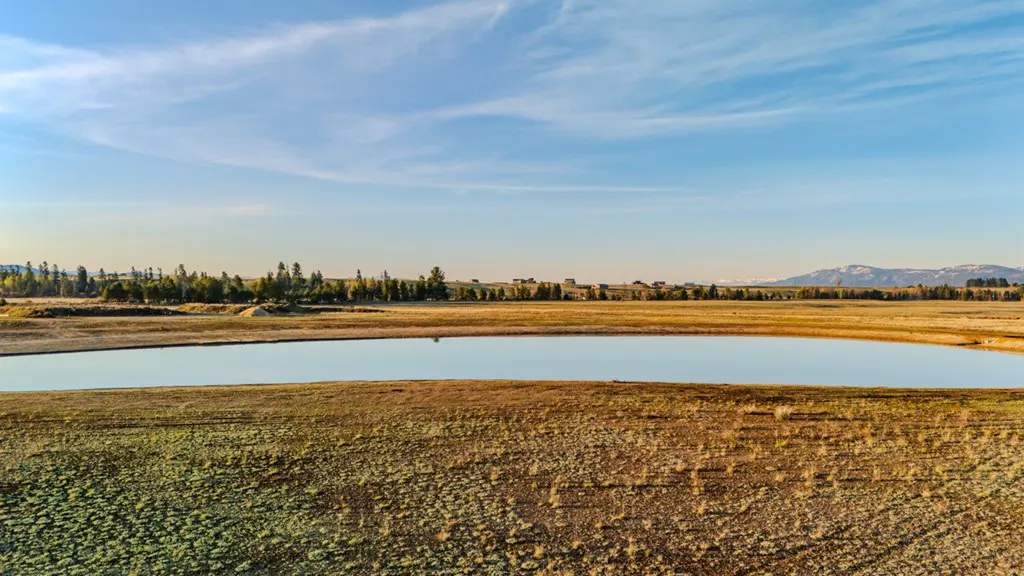Landscape view of a pond surrounded by fields under a blue sky with wispy clouds. Trees and distant mountains are visible.