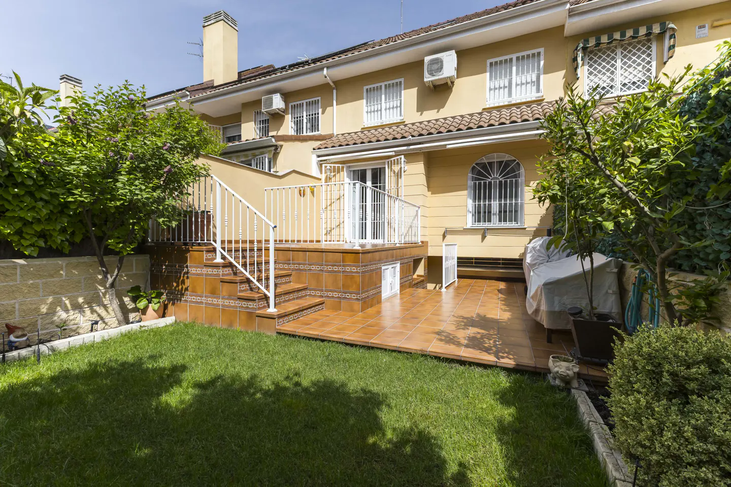 Backyard view of a two-story, yellow house with a tiled patio, white railing, green lawn, and trees under a blue sky.