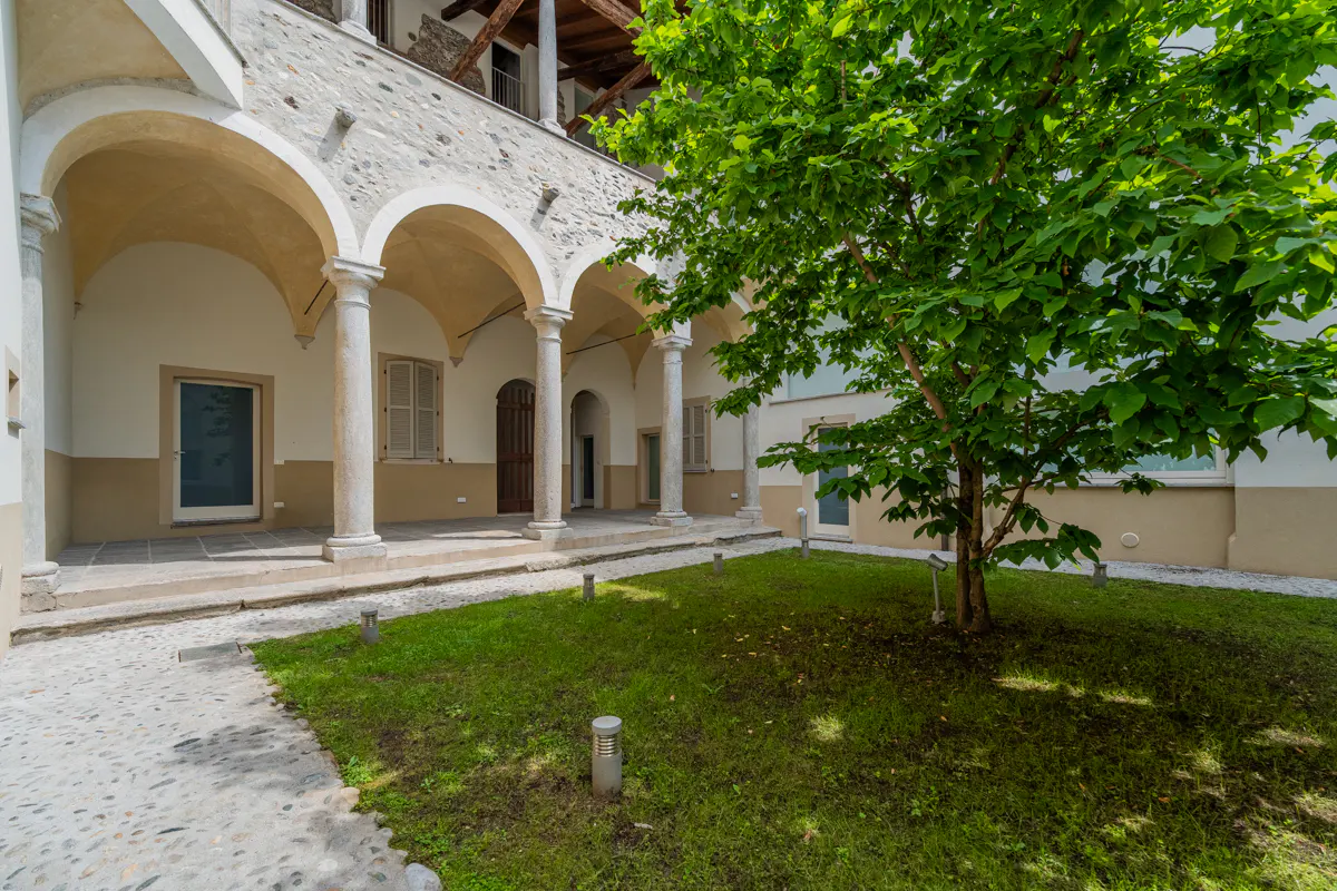 Courtyard with arched walkway, stone columns, and green lawn with a tree.