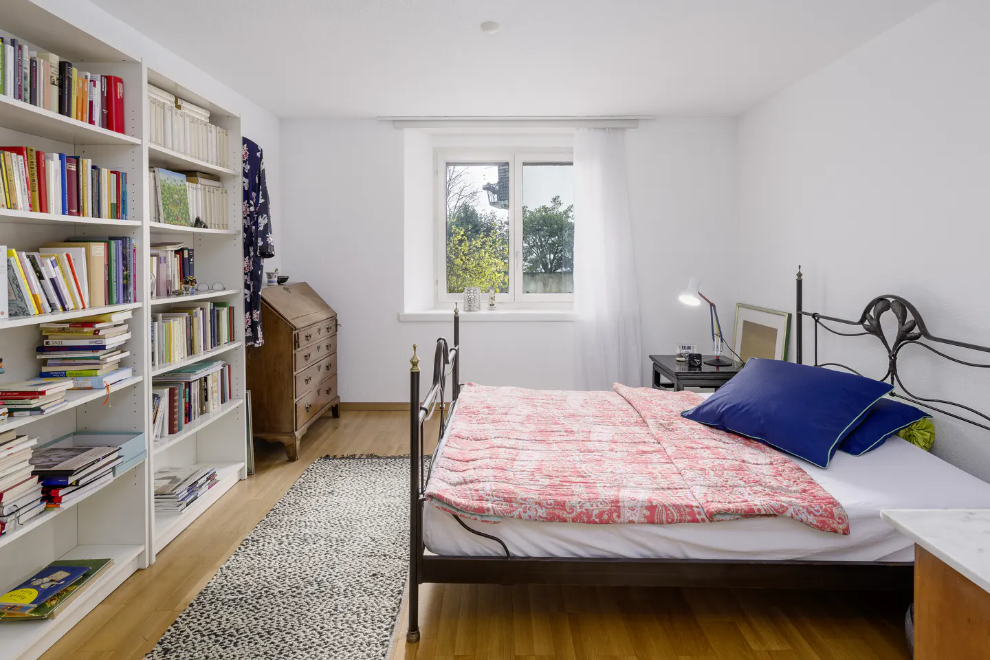 Bedroom with white walls, hardwood floors, and a black metal bed frame. A large bookcase filled with books is on the left. A window is centered on the back wall.