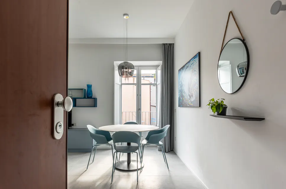 A partially open door reveals a dining area with a round table and blue chairs. A mirror and artwork adorn the white wall.