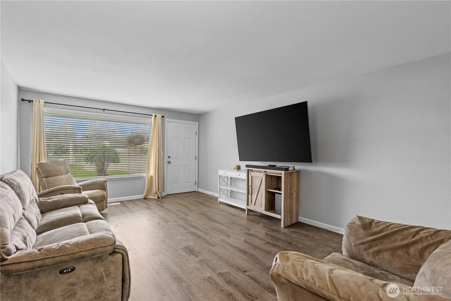 Living room with gray walls, wood floors, and tan furniture. A large TV is mounted above a rustic wood cabinet. A window with blinds and curtains is on the left.