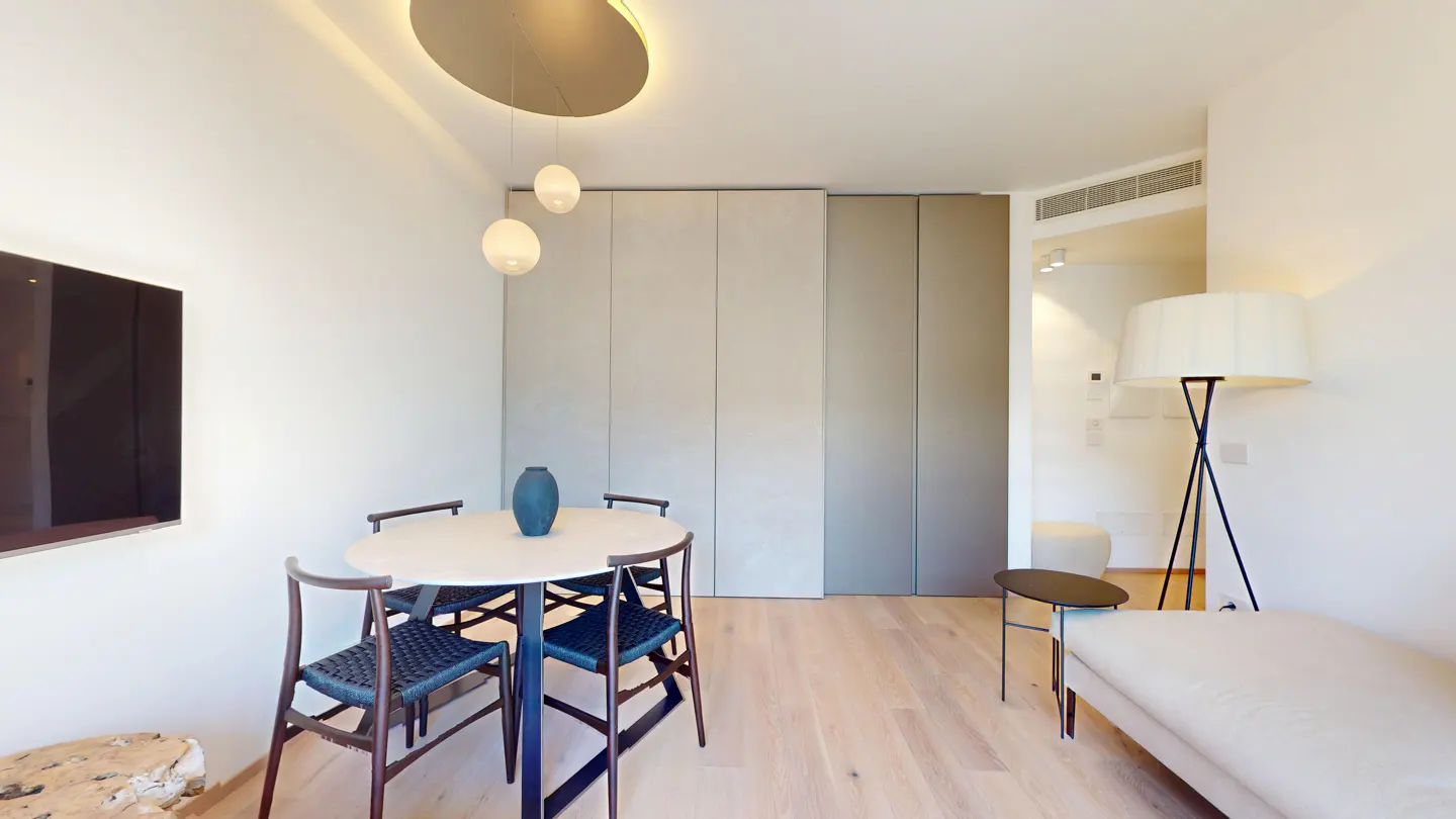 Modern dining area with a round table, four dark chairs, a blue vase, and a floor lamp next to a sofa on light wood floors.