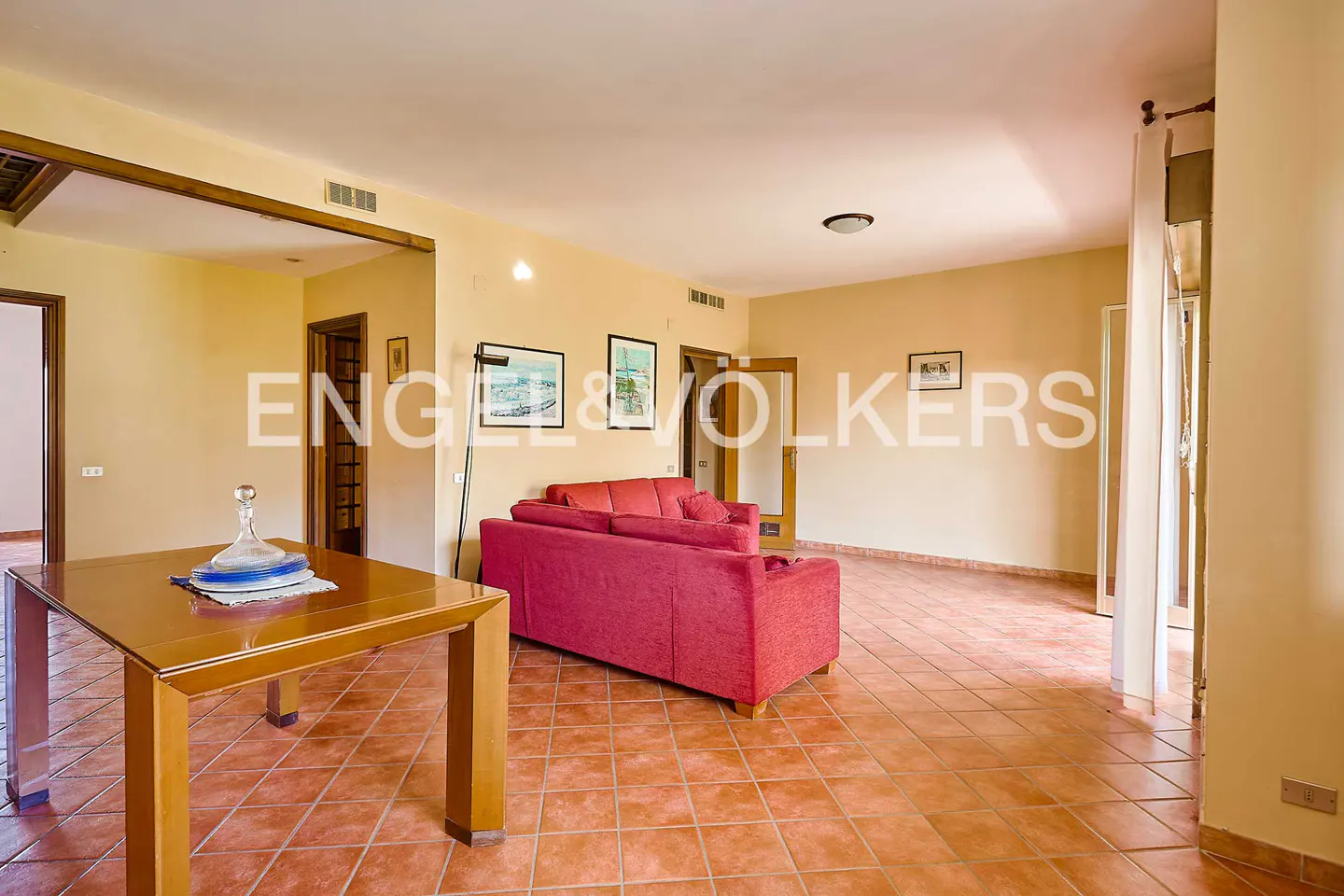 Spacious living room with terracotta tile floors, a red sofa, and a wooden dining table with a glass decanter.