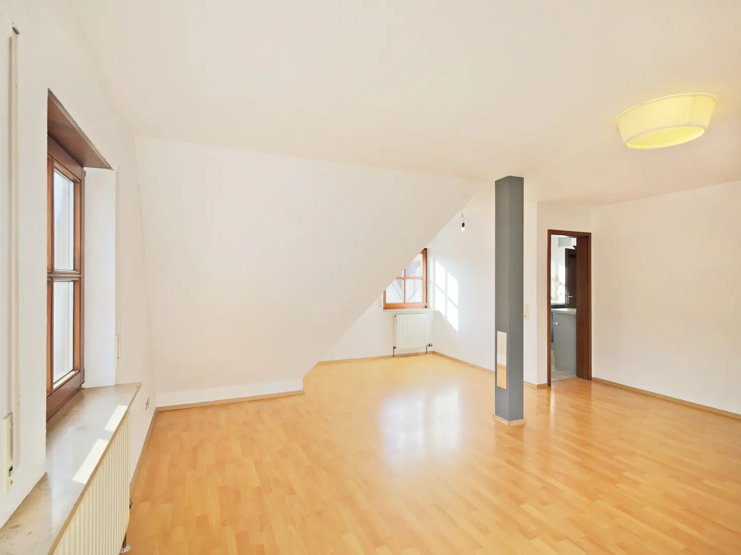 Bright, empty room with wood floors, white walls, and natural light from two windows. A gray pillar stands near an open doorway.