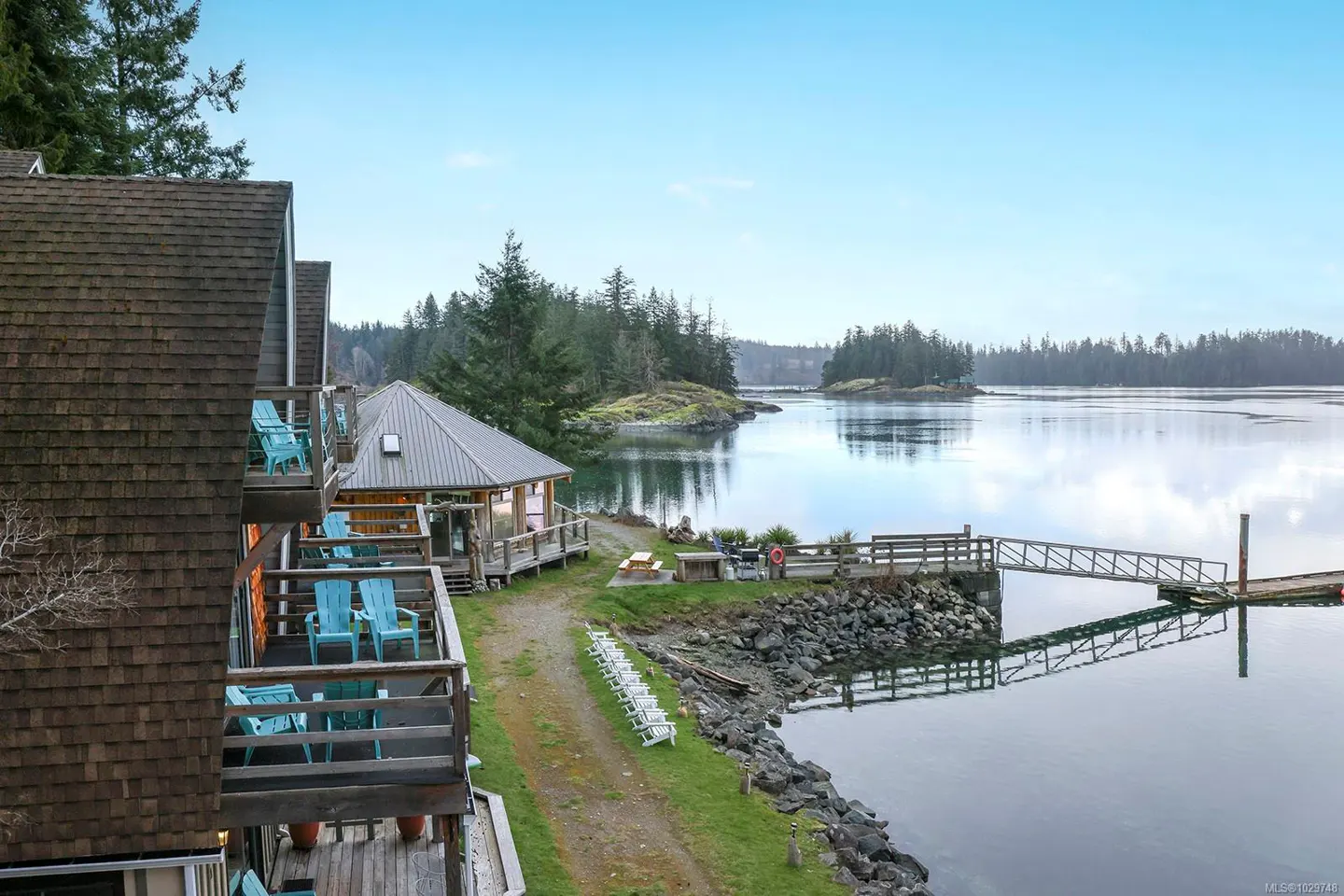 Waterfront property with a dock, a small house, and balconies with blue chairs. The sky is blue and the water is calm.