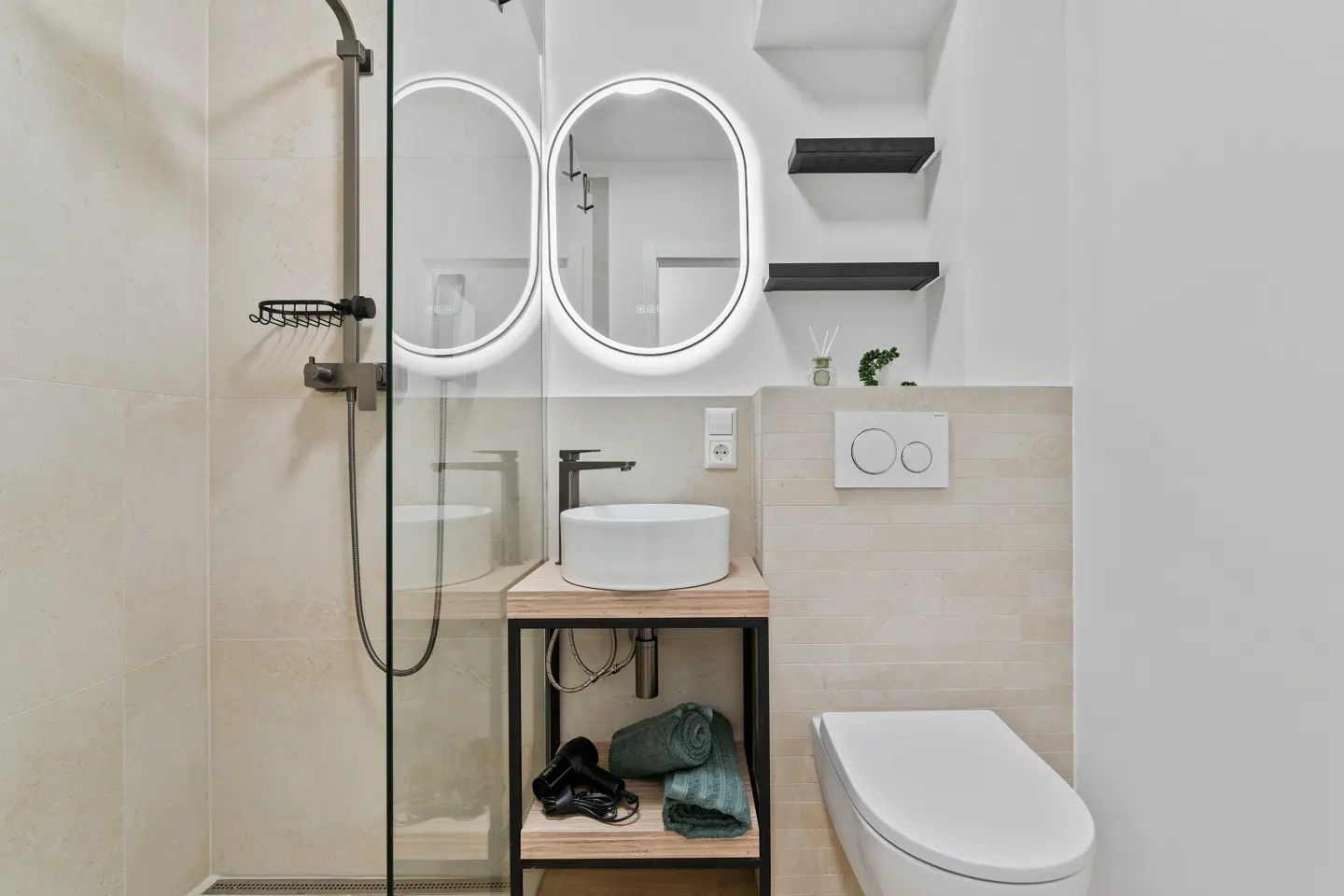 Modern bathroom with a glass shower, white sink on a wooden stand, and a toilet. Two oval mirrors with lights above the sink.