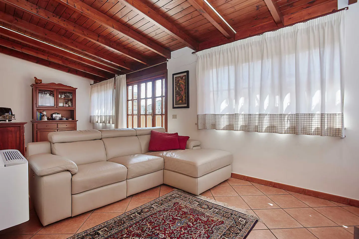 Living room with a beige leather sofa, red pillows, and a patterned rug on terracotta tile. White curtains hang near a wood-beamed ceiling.
