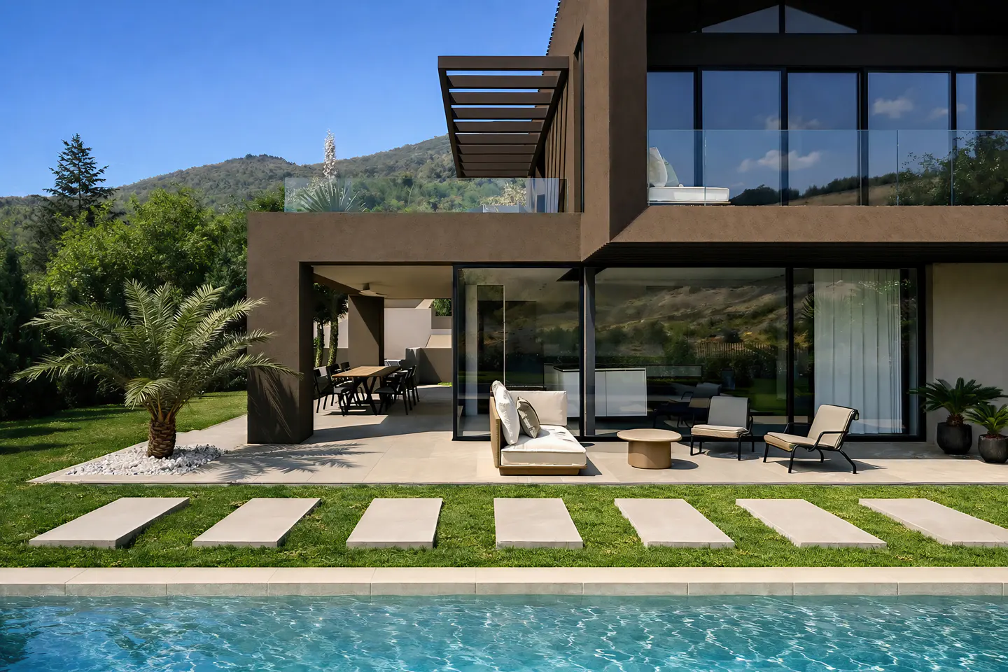 Modern brown house with a pool, patio furniture, and a dining table. Green grass and trees surround the house.
