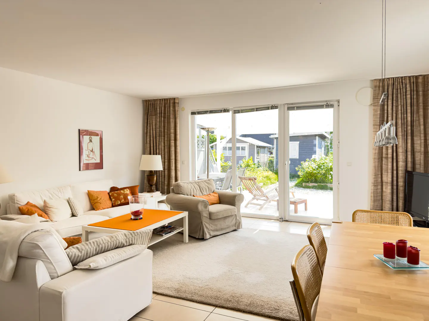 Bright living room with white sofa, beige armchair, and wooden table. Large windows overlook a green yard with a small building.