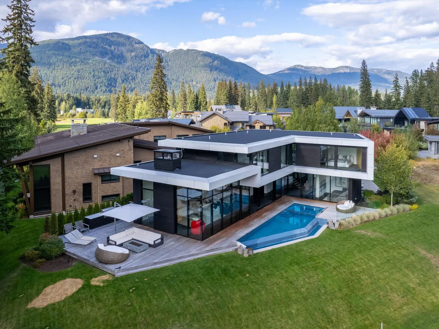 Modern black and white house with a pool and wooden deck, surrounded by green grass and trees, with mountains in the background.