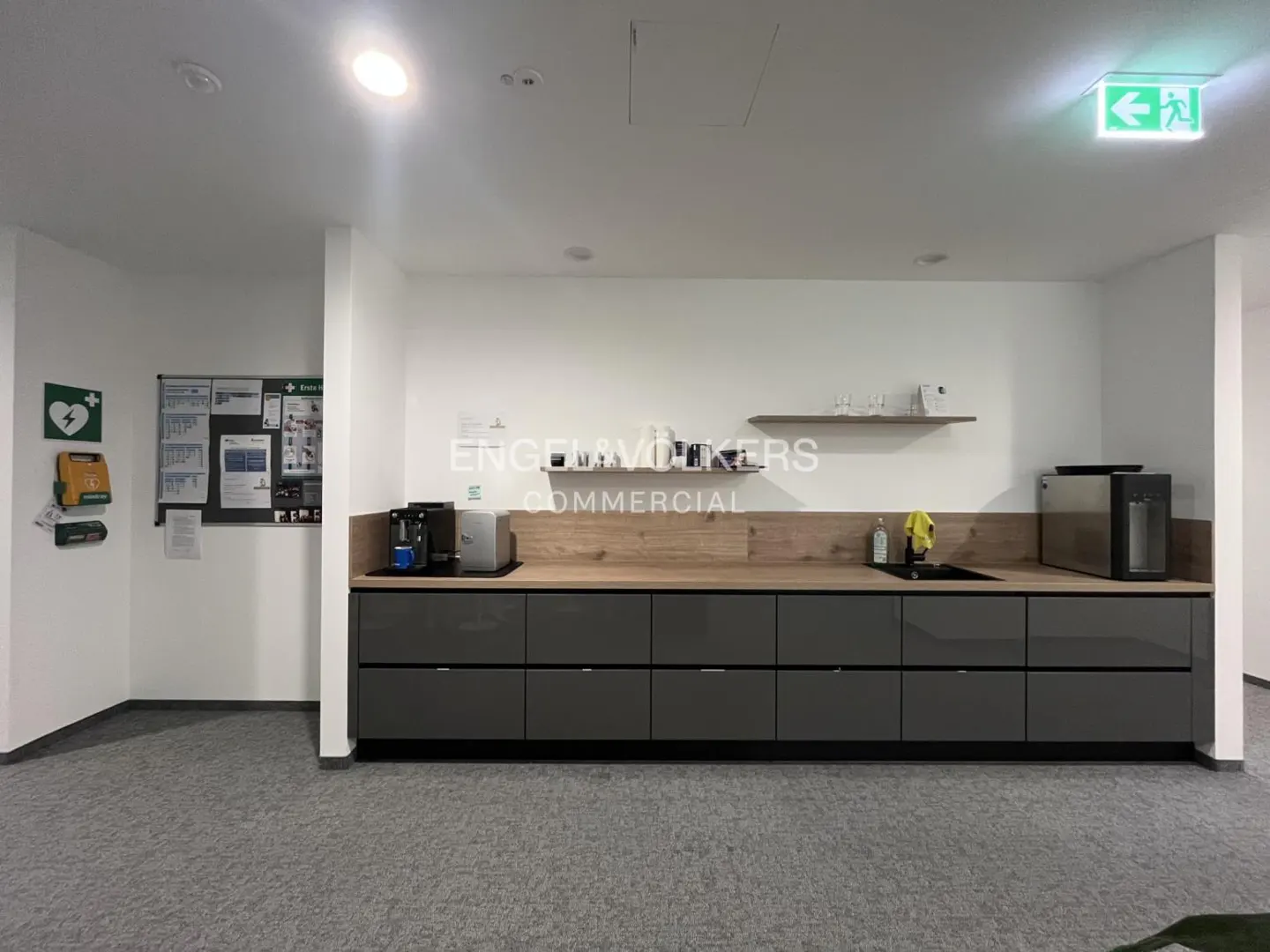 Office kitchenette with gray cabinets, wood countertop, sink, coffee maker, and water dispenser. Emergency exit sign visible.