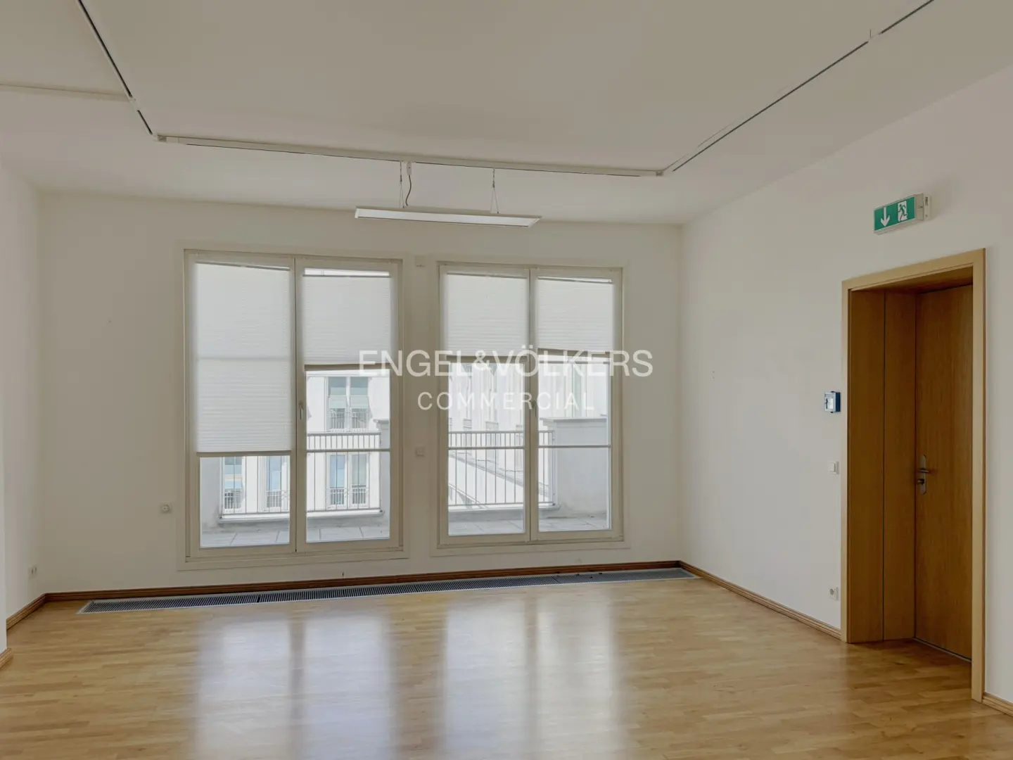Bright, empty room with wood floors, white walls, and two large windows with blinds. A wooden door with an exit sign is on the right.