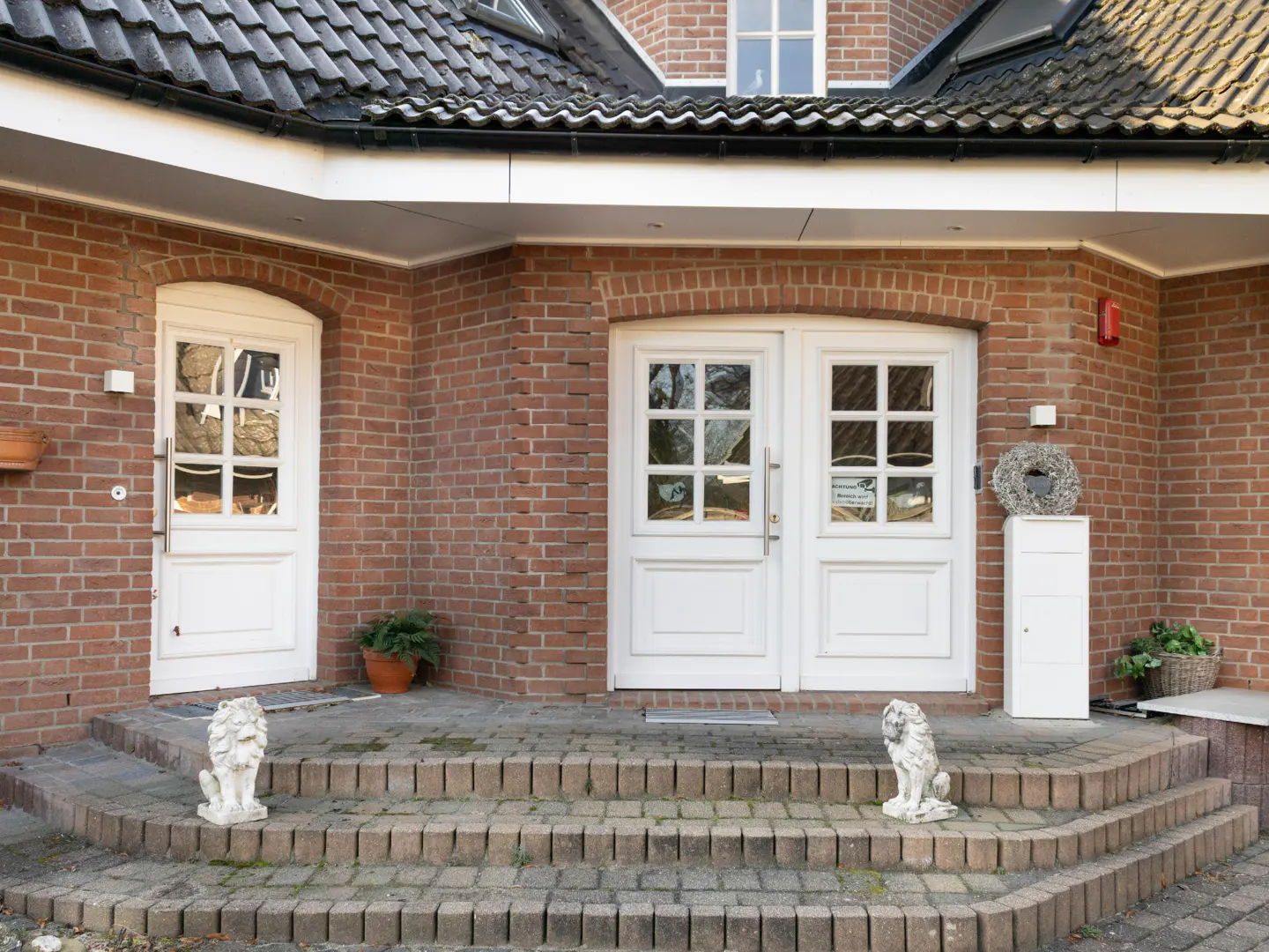 Brick house entrance with white doors, lion statues, and stone steps. A wreath hangs near the double doors.