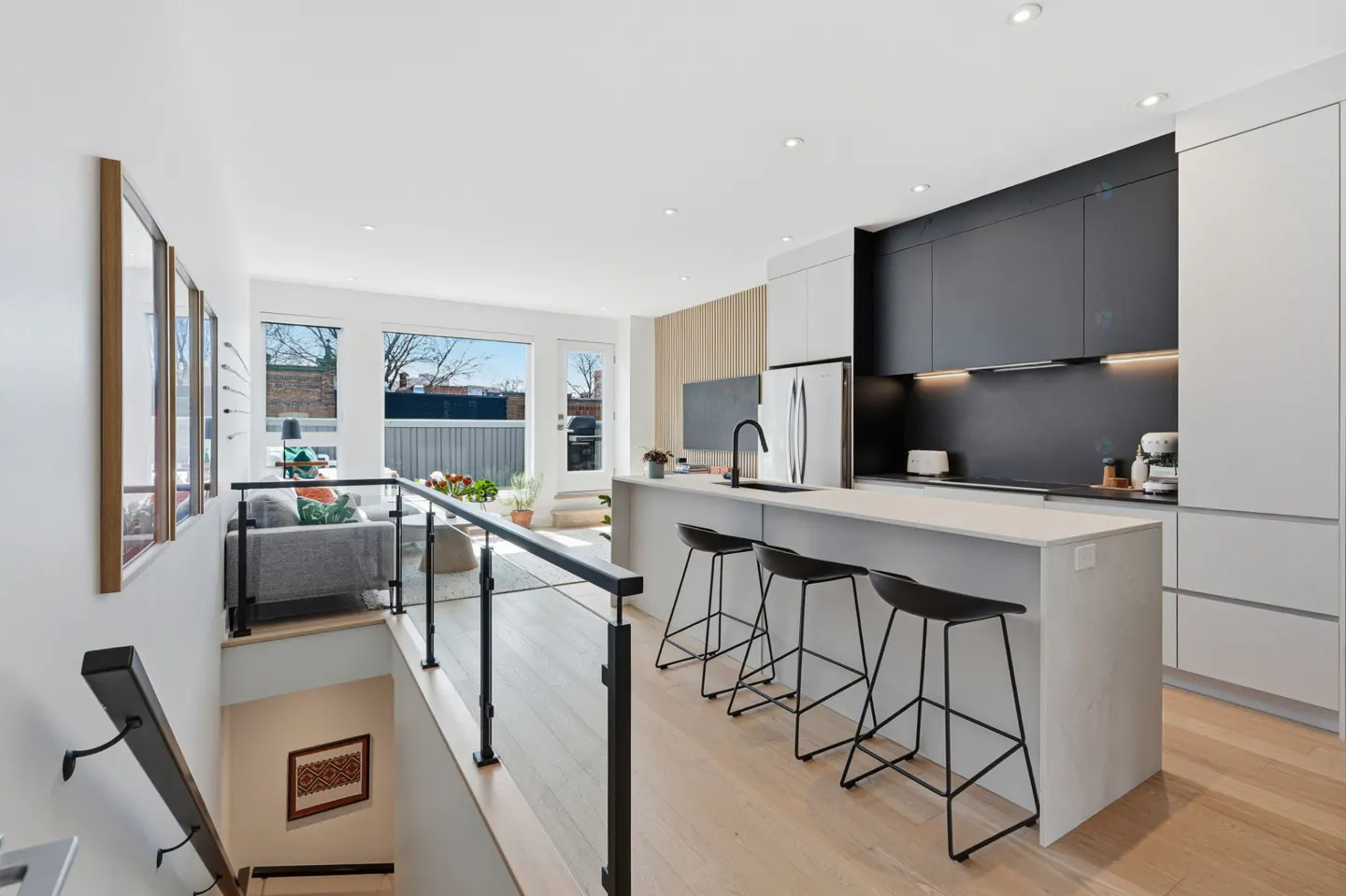 Bright, modern kitchen with black and white cabinets, island with stools, and a view to the living room. Stairs lead down to another level.