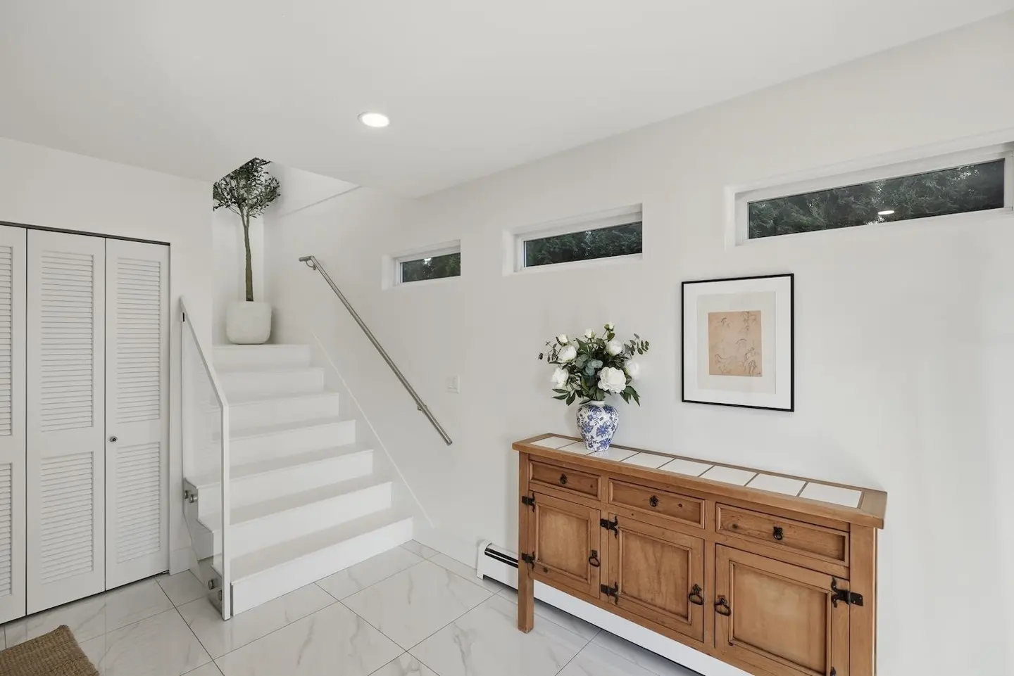 Bright foyer with white walls, marble floors, and a wooden cabinet topped with a blue and white vase of white flowers. White stairs with a silver handrail lead up.