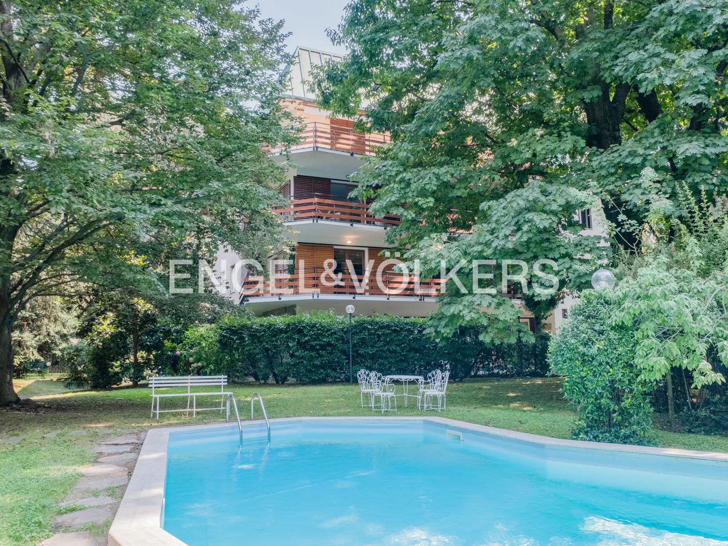 Exterior view of a multi-story building with balconies, a pool, and lush greenery. A table and chairs sit on the lawn.