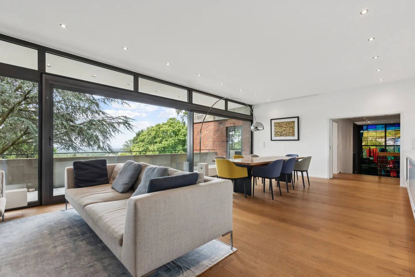 Modern living room with a beige sofa, dining area, hardwood floors, and floor-to-ceiling windows overlooking trees.