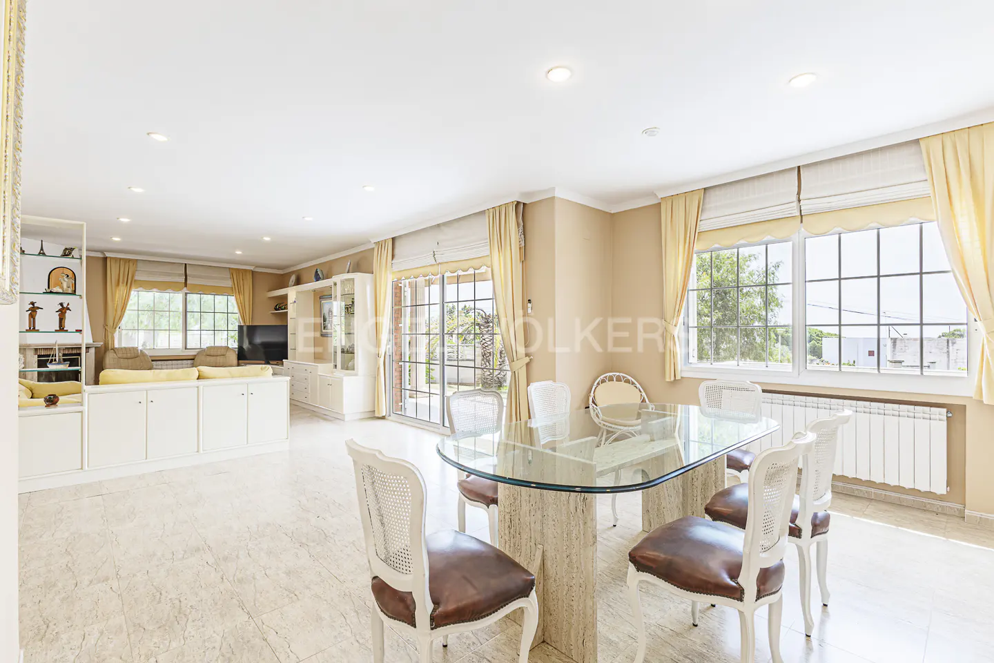 Bright, open-plan dining and living room with a glass-topped table, brown leather chairs, and large windows with yellow curtains.