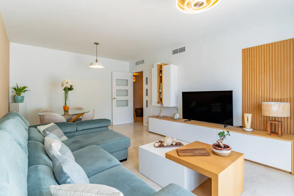 Bright living room with a blue sectional sofa, white walls, and a modern entertainment center with a large TV. A wood and white coffee table sits in front.