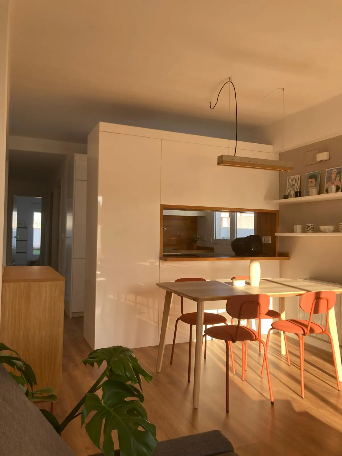 Sunlit modern dining area with a light wood table, orange chairs, and white cabinets. A monstera plant is in the foreground.