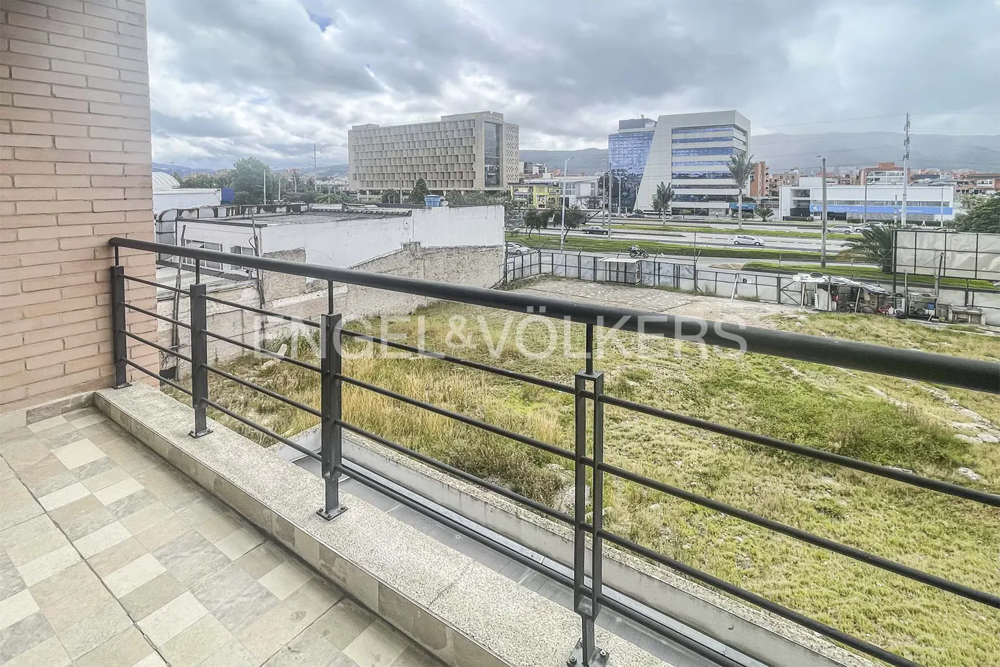 Balcony view with black metal railing overlooking a grassy lot and city buildings under a cloudy sky. Brick wall on the left.