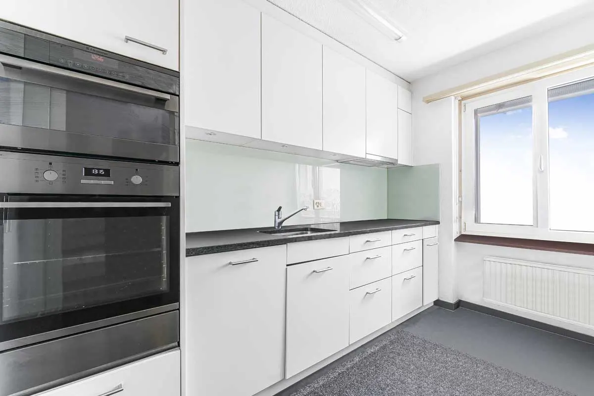 Bright kitchen with white cabinets, dark countertops, and stainless steel oven. A window shows a blue sky.