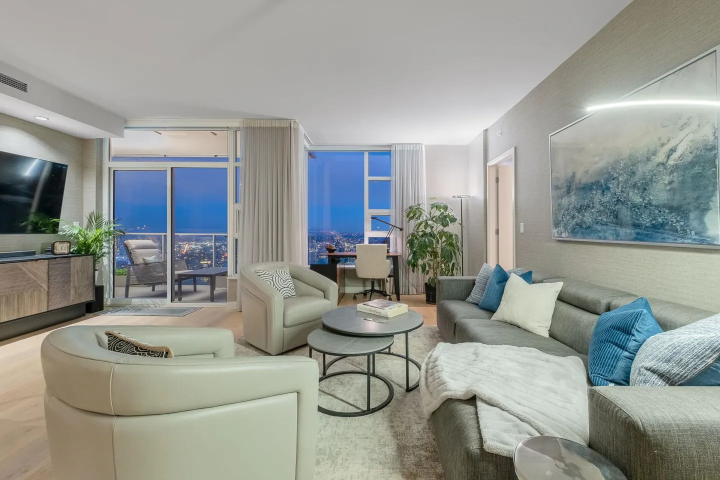 A modern living room with gray sofa, light armchairs, and a city view at dusk. A desk area is visible by the window.