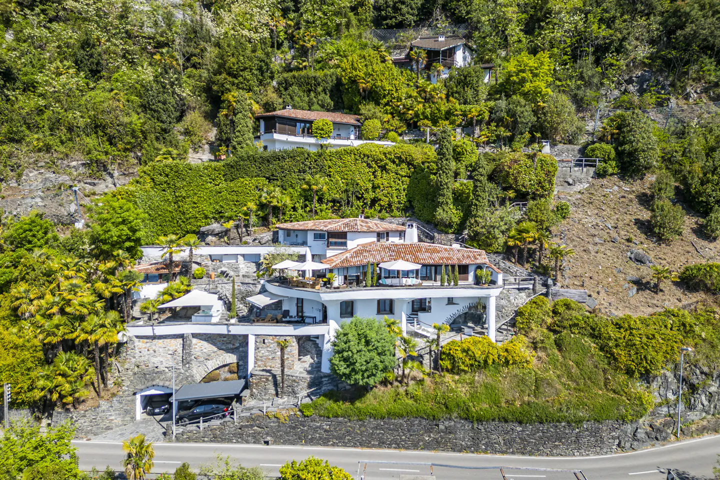 Aerial view of a white villa with a red tile roof nestled on a lush green hillside. Stone archways support the structure.