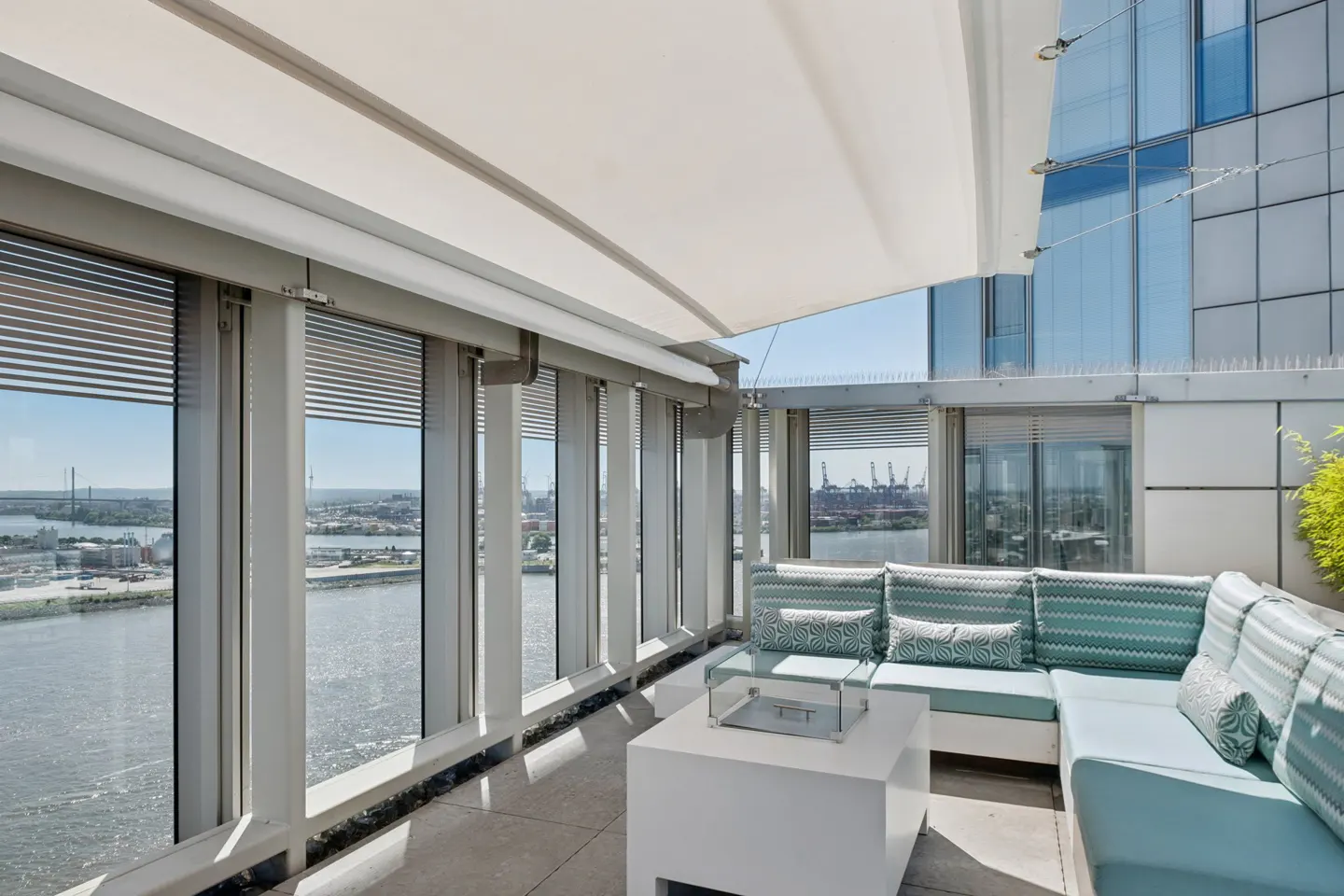 Modern balcony with a white sectional sofa, glass fire pit, and a retractable awning overlooking a harbor and city skyline.