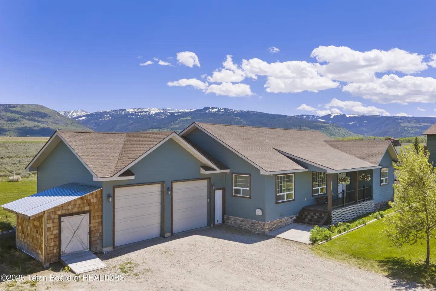 Exterior of a blue house with a brown roof, two-car garage, and a shed. Mountains and a blue sky with clouds are in the background.