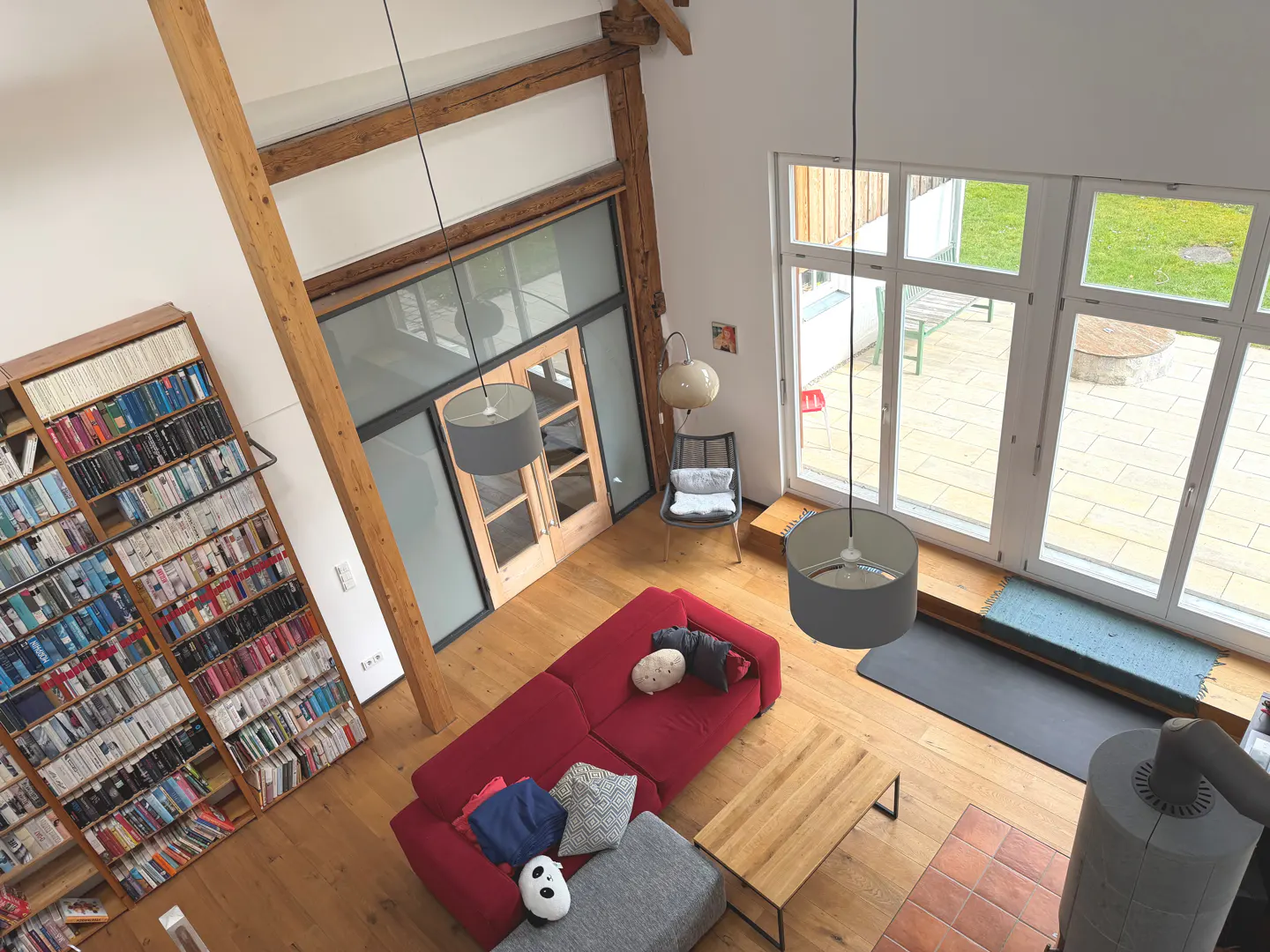 High-angle view of a living room with a red sofa, wooden floors, and a tall bookshelf filled with books. Large windows overlook a green lawn.