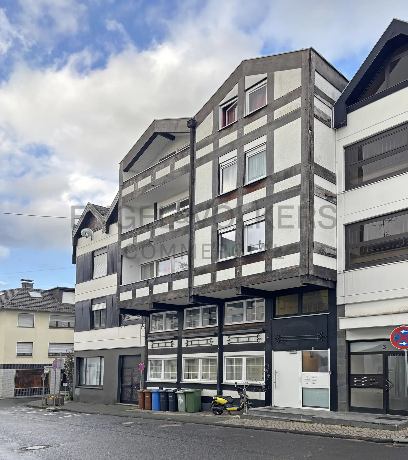 A three-story building with white walls and dark brown timber framing, under a cloudy sky. A yellow scooter is parked in front.