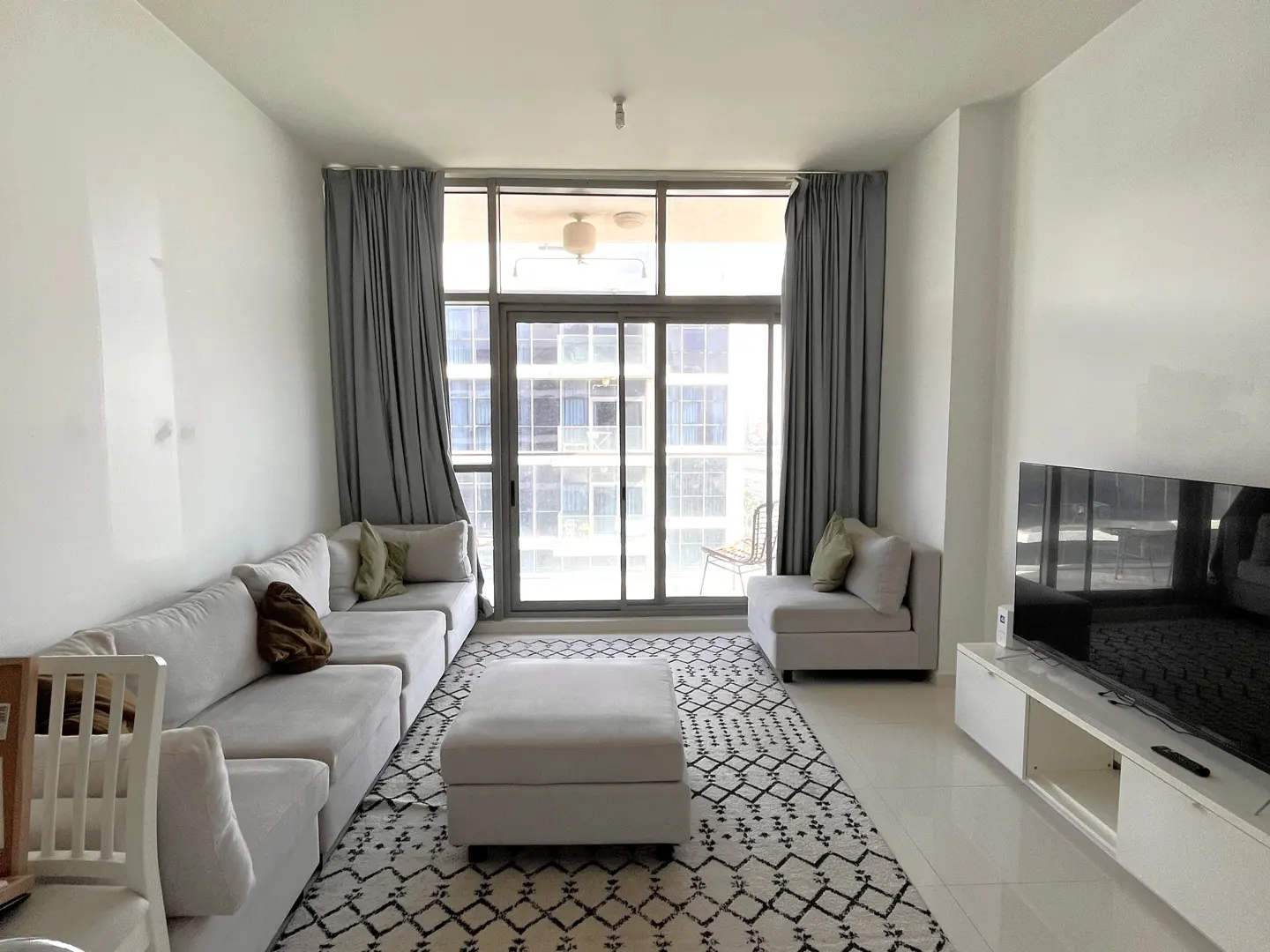 Bright living room with white walls, tile floor, and a large window with gray curtains. A white sectional sofa, ottoman, and patterned rug fill the space.
