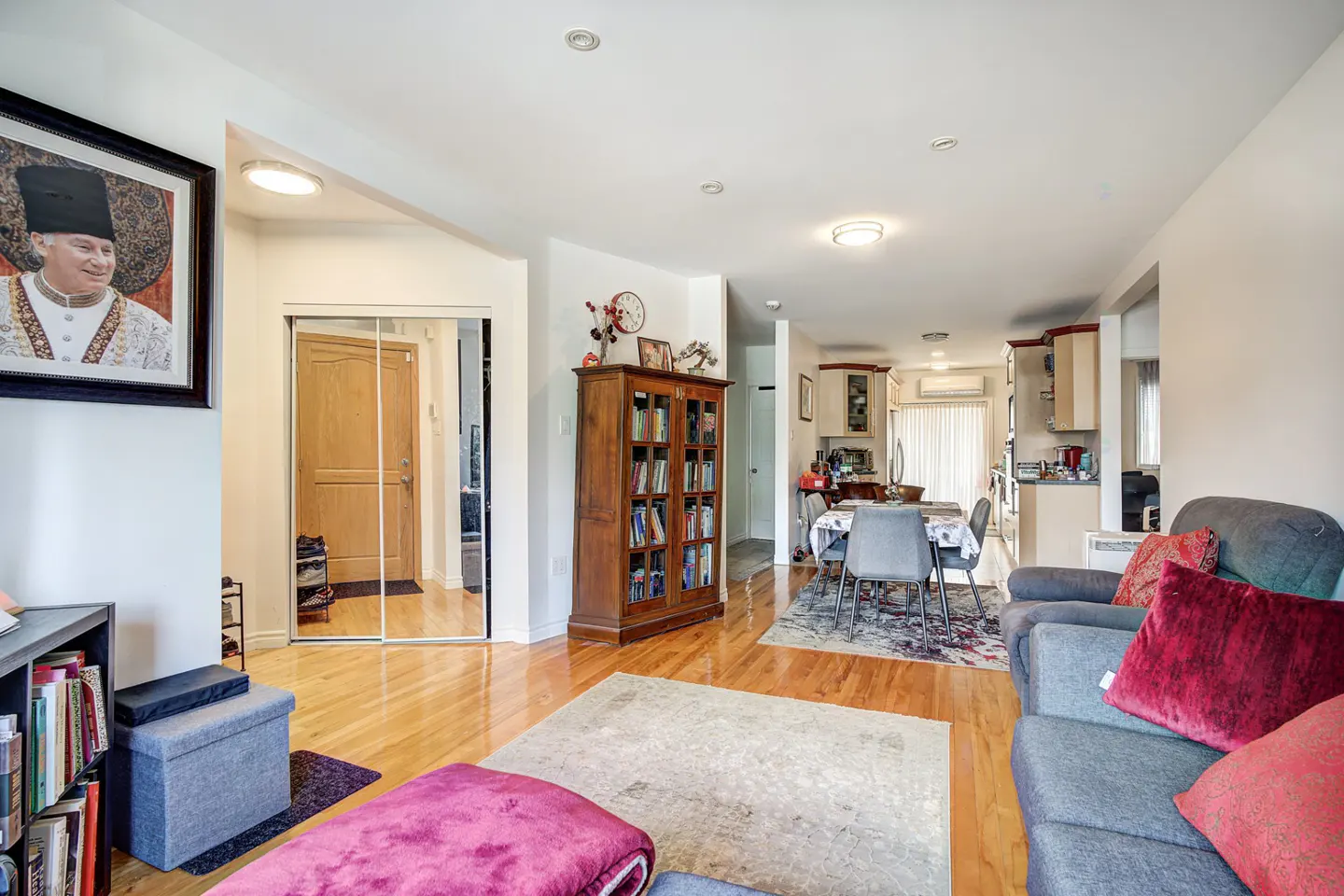 A bright living room with hardwood floors, a gray sofa with red pillows, and a dining area leading to the kitchen. A framed portrait hangs on the wall.