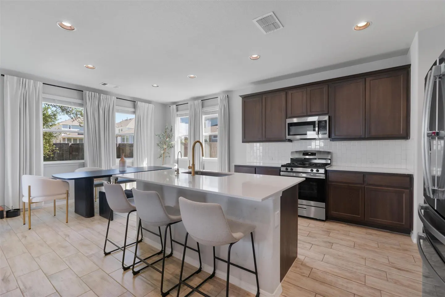 Bright kitchen with dark wood cabinets, stainless steel appliances, and a white countertop island with three stools. A dining table sits near the windows.
