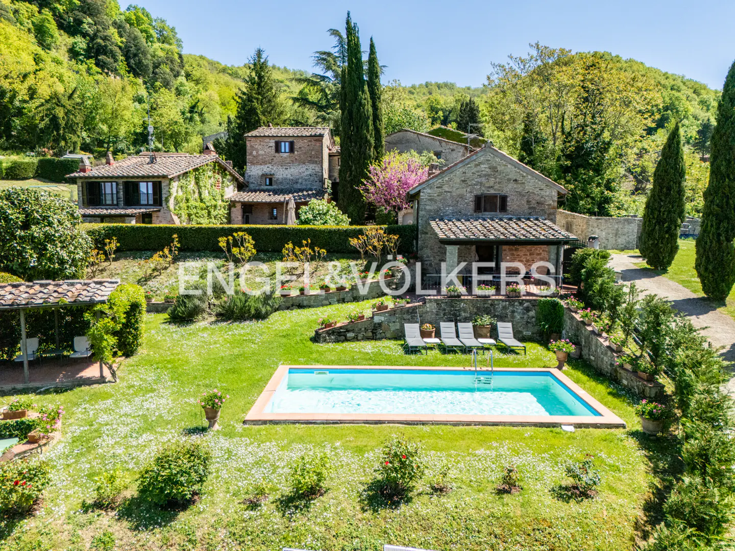 Aerial view of a stone house with a pool, lounge chairs, and green lawn in a lush, hilly landscape.