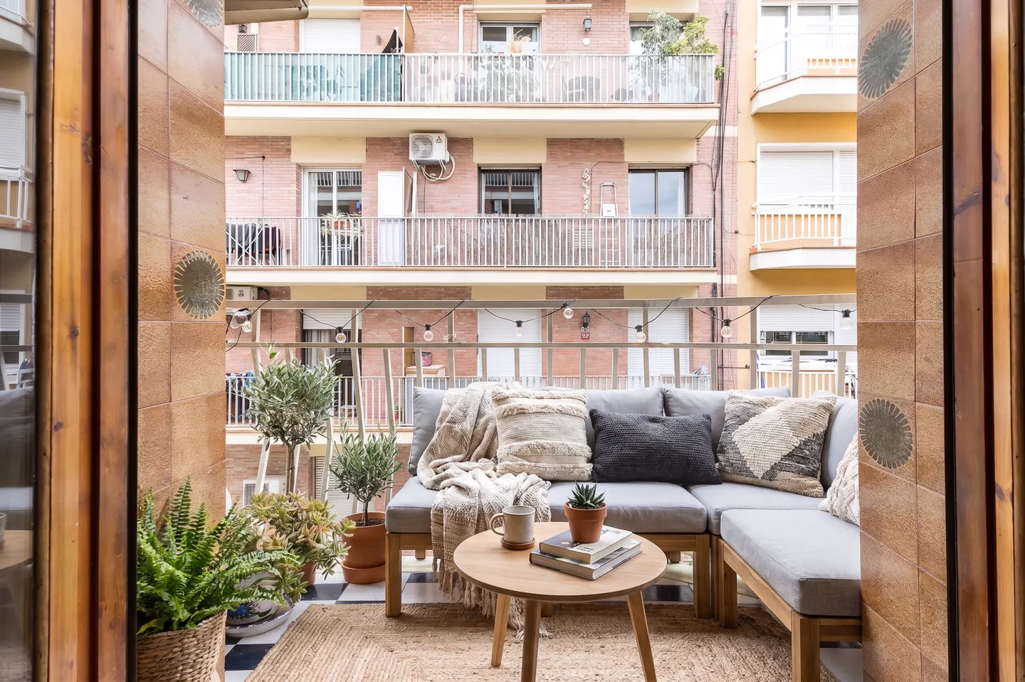 A cozy balcony with a gray sectional sofa, throw pillows, a round wooden table, and potted plants, with string lights overhead.