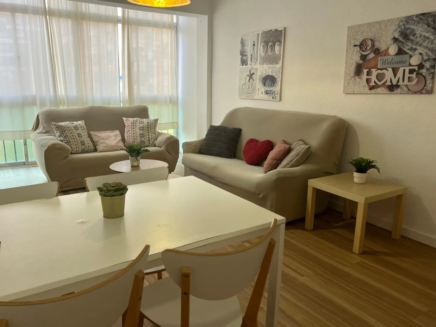 Bright living room with two beige sofas, a white dining table, and wall art, featuring light wood floors and sheer curtains.