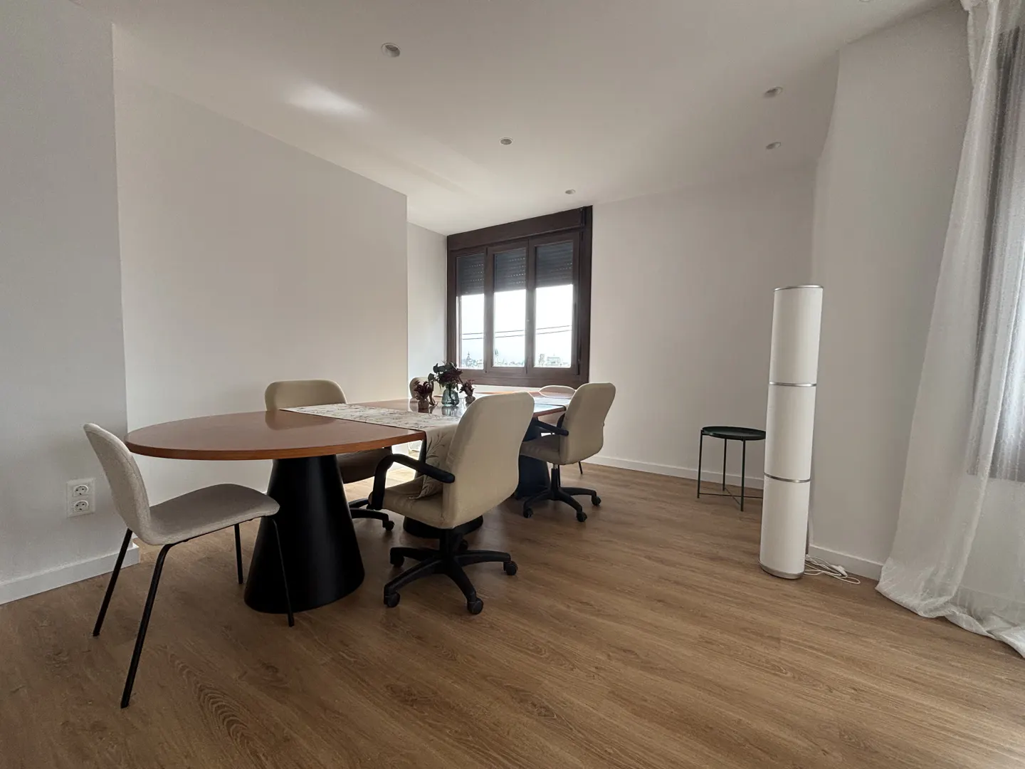 Bright dining room with wood floors, a round wood table, and beige chairs. A window with dark blinds is in the background.