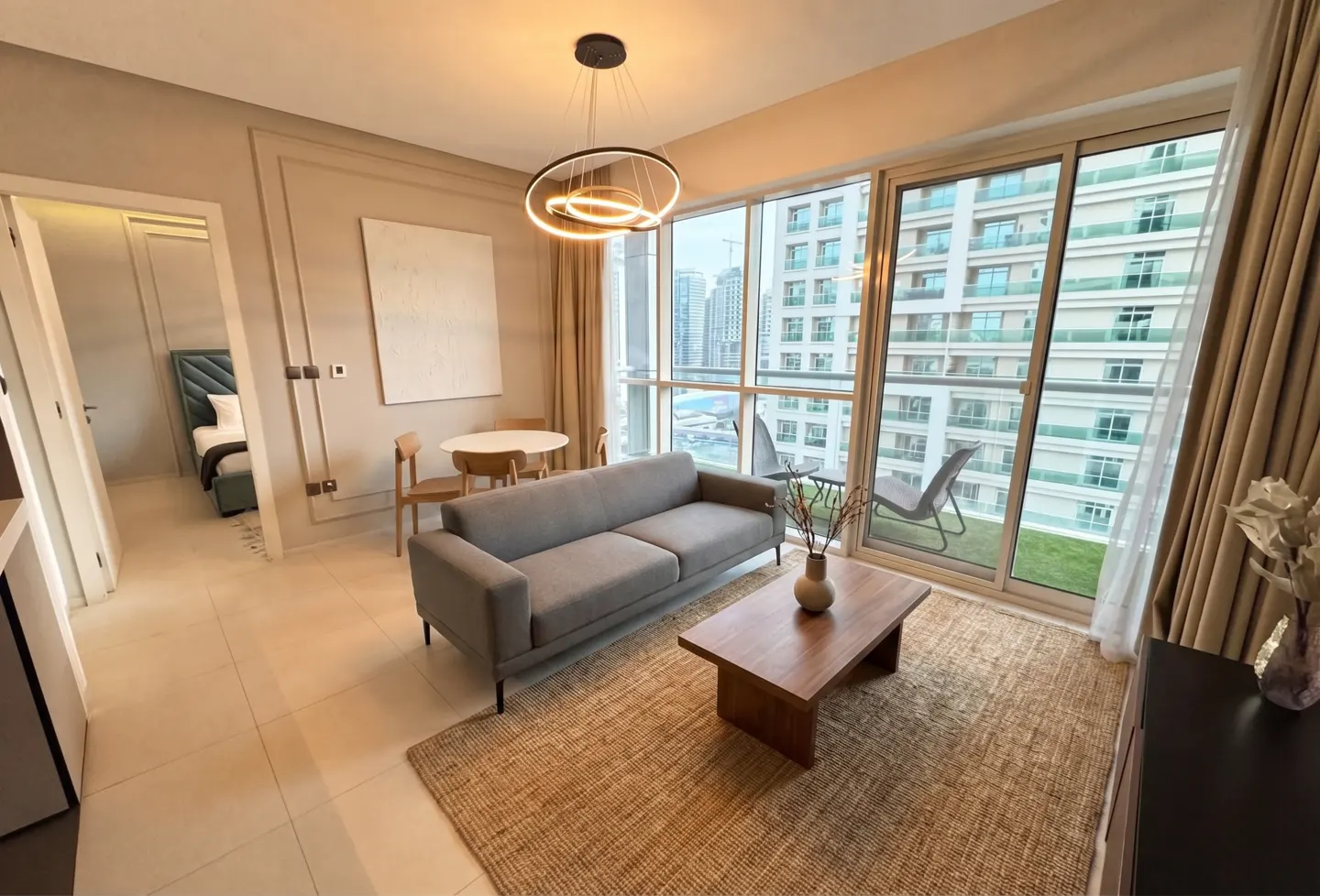 A modern apartment living room with a gray sofa, wood table, and city view. A bedroom is visible through an open door.