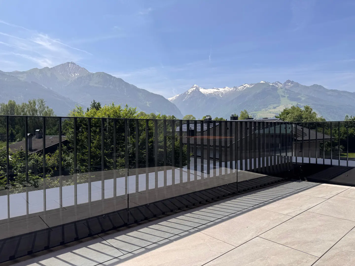Balcony view with a black metal railing, trees, houses, and snow-capped mountains against a blue sky.